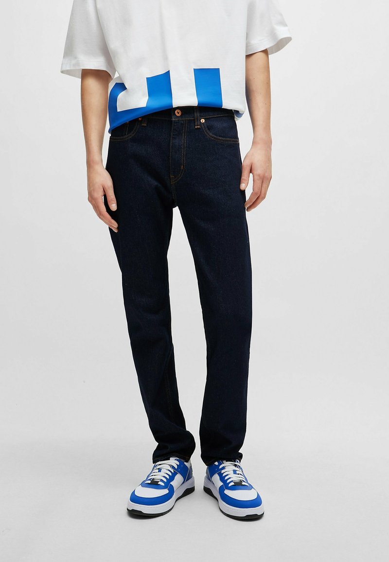 Personne portant un jean bleu foncé, un t-shirt blanc avec des motifs bleus et des baskets blanches avec des touches de bleu, debout devant un fond uni.