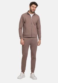 Veste zippée marron avec poches avant, associée à un pantalon de jogging ajusté assorti. Les deux sont fabriqués dans un tissu doux, stylisés avec des poignets et des ourlets côtelés.