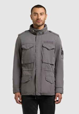 Férfi, szürke, több zsebes zsebekkel és magas gallérral rendelkező munkadzsekit visel, egyszínű világos háttér előtt áll.