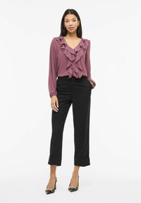 Lilla bluse med volanger, V-hals og lange ermer, kombinert med svarte skreddersydde bukser og svarte pumps med gullaksenter.