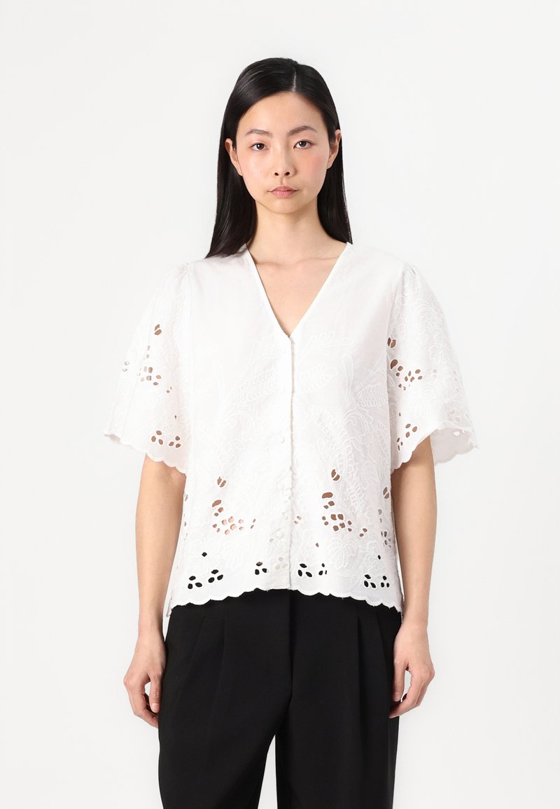 White, short-sleeve blouse featuring intricate floral embroidery and cut-out patterns along the hem, with a V-neckline and button closure.