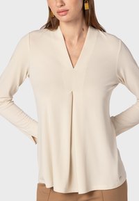 Luftige, langärmlige Bluse in hellbeige mit V-Ausschnitt und Faltendetail an der Vorderseite. Weicher Stoff, minimalistisches Design mit dezenter Markenkennzeichnung.