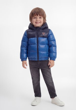 Bambino sorridente che indossa una giacca imbottita blu e blu navy, jeans neri e sneaker bianche, in piedi contro uno sfondo bianco.