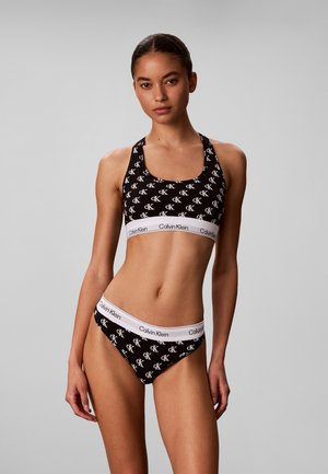 Femme portant un soutien-gorge de sport Calvin Klein noir et des sous-vêtements assortis avec des bandes élastiques blanches siglées, debout devant un fond gris uni.