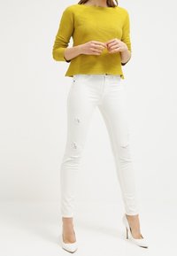 Haut à manches longues jaune côtelé associé à un jean skinny blanc avec des détails déchirés et des patches usés, complété par des talons hauts blancs.