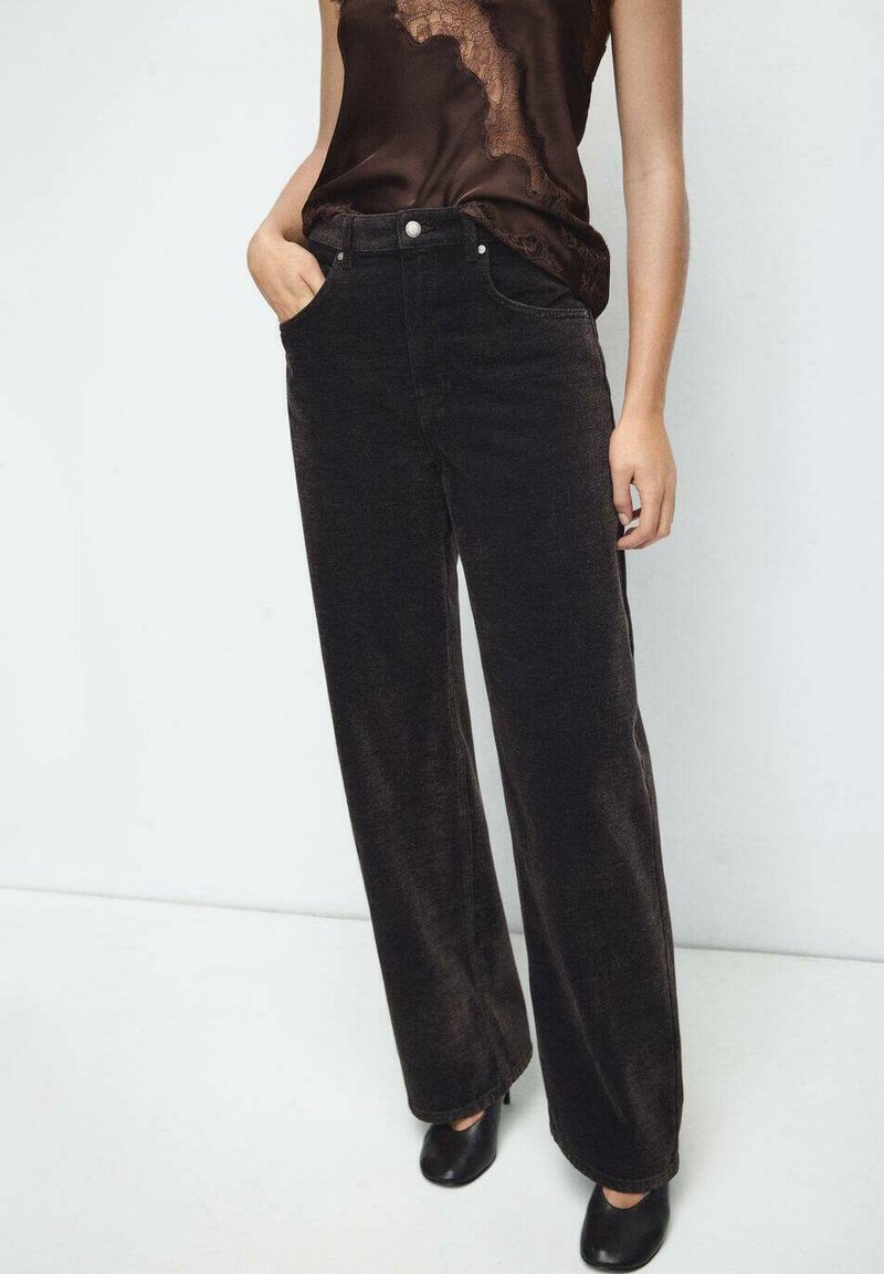 Pantalones anchos de pana negra con bolsillos delanteros, cierre con cremallera y botón. Combinados con una blusa de satén oscura y tacones negros. Textura suave.