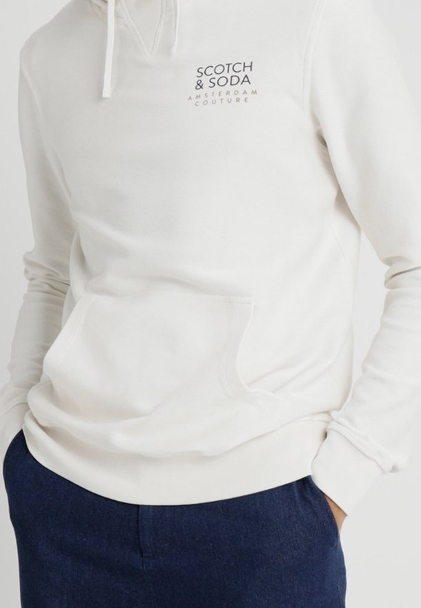 Sweat à capuche blanc avec poche avant et cordons, avec le logo "Scotch & Soda Amsterdam Couture" sur la poitrine, porté avec un pantalon bleu foncé.