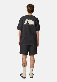 Czarna bawełniana koszulka z białym wzorem na plecach, zestawiona z czarnymi szortami; widoczne tatuaże i jasne skarpetki.