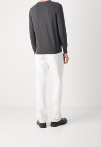 Homme portant un haut à manches longues gris foncé, un pantalon blanc et des chaussures noires, debout dos à la caméra sur fond blanc.