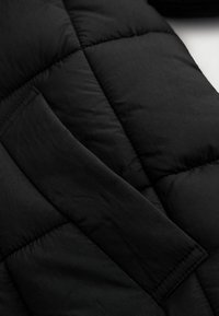 Czarna pikowana kurtka puchowa z miękką teksturą, geometrycznym przeszyciem i ukrytym detalem kieszeni na gładkiej powierzchni tkaniny.