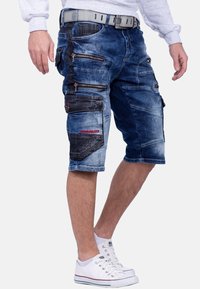 Shorts cargo en denim bleu avec plusieurs poches zippées et des accents en panneau. Finition droite avec ourlet roulé, associés à des baskets blanches.