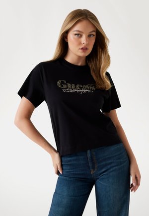 Black cotton t-shirt with a rounded neckline and short sleeves, featuring a shiny gold "Guess" logo and "Nineteen Eighty One" text detail.