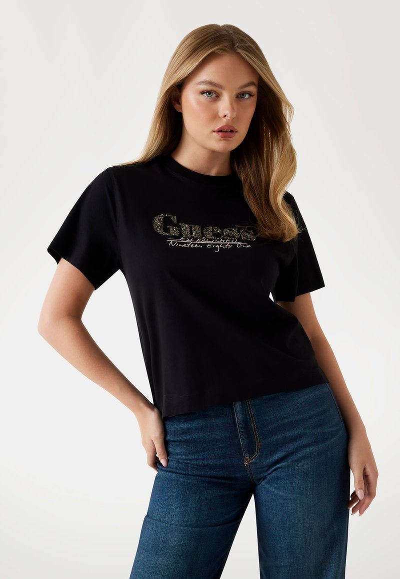 Must mõnusa ümara kaelusega must puuvillane T-särk lühikeste varrukatega, millel on särav kuldne "Guess" logo ja "Nineteen Eighty One" tekstiline detail.