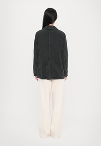 Giubbotto in denim scuro con colletto e cuciture verticali, abbinato a pantaloni bianchi larghi. Il tessuto appare texturizzato con una finitura leggermente sbiadita.