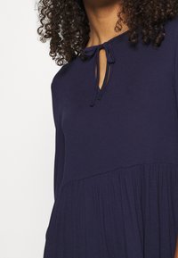 Kvinna med lockigt hår som bär en mörkblå klänning med en nyckelhålsutskärning knuten med en rosett och detaljer med veckad kjol.