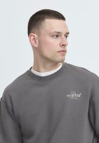 Grå hettegenser med rund hals, ribbede mansjetter og en liten hvit brodert logo på brystet. Myk tekstur og avslappet passform.