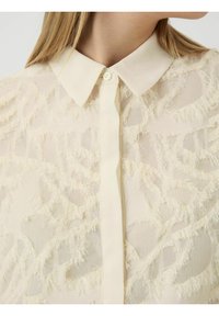 Kermainen läpikuultava paita, jossa on teksturoitu, risainen muotoilu; mukana klassinen kaulus ja napitus edessä.