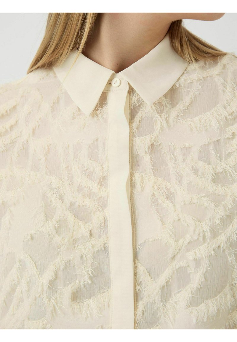 Translucentes Cream-Shirt mit einem strukturierten, fransigen Design; verfügt über einen klassischen Kragen und eine Knopfleiste auf der Vorderseite.