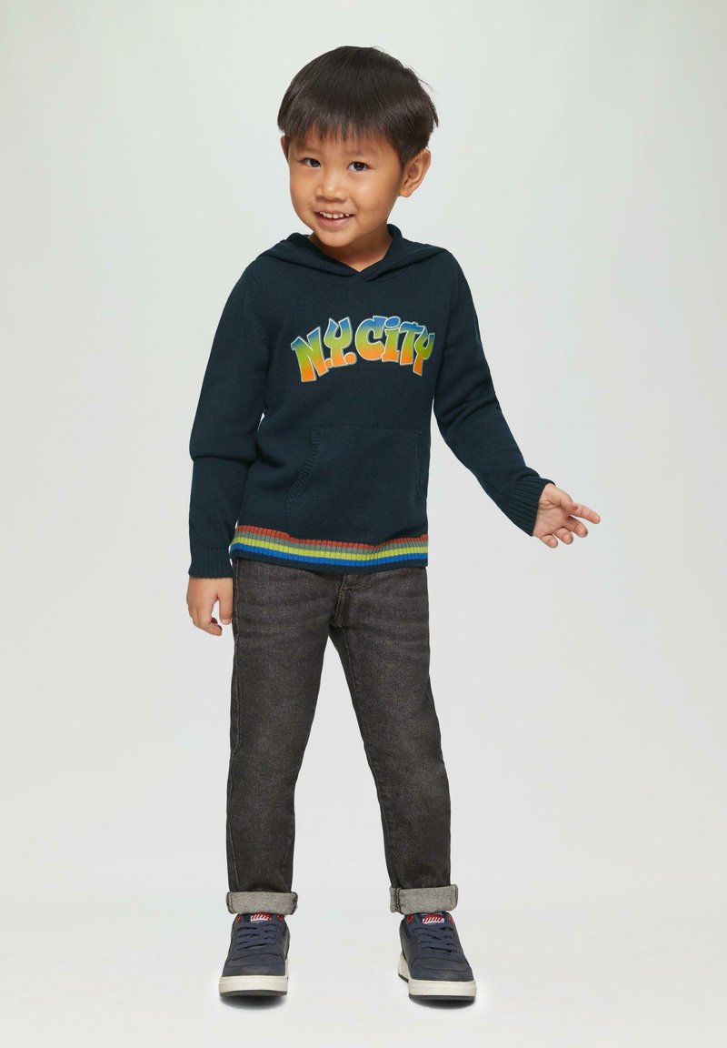 Navy gebreide hoodie met "NY CITY" graphic in gradient kleuren, kangoeroezak, gestreepte ribs aan de onderkant. Gecombineerd met donkere jeans en sneakers.