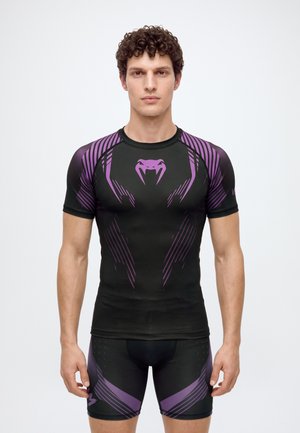 Un mannequin masculin se tient devant un fond blanc, vêtu d’un haut de compression noir et violet et d’un short assorti.
