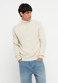 Crèmewit coltruit in een gestructureerde brei, met lange mouwen en een geribbelde zoom, gecombineerd met lichtblauwe jeans.