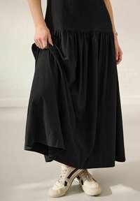 Rochie maxi neagră cu detalii de tier strâns la tiv, purtată cu adidași bej cu dungi negre. Textură netedă, design fluid.