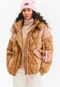Brązowa pikowana kurtka z różowymi akcentami, z teksturowanymi panelami, kieszeniami po bokach oraz suwakami z chwostami. Noszona z beżowym golfem.