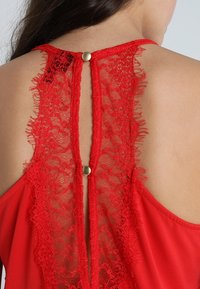 Rood kledingstuk met een rug van kant, voorzien van een keyhole-uitsnijding en goudkleurige knoopdetails. Gladde stof met een vloeiende silhouet.