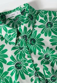 Camicia a maniche lunghe con motivo floreale verde e bianco, collo a camicia e chiusura con bottoni, caratterizzata da grandi margherite verdi con centri neri.