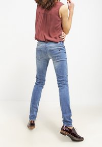 Jeans ajustados de color azul con un ligero estiramiento, de talle medio, y detalles en los bolsillos, combinados con una blusa sin mangas de color marrón y zapatos de color marrón oscuro.