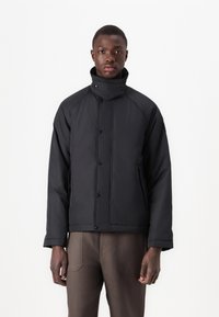 Black padded jacket with a high collar, snap-button closure, and side pockets. Features a smooth texture and minimalistic design.