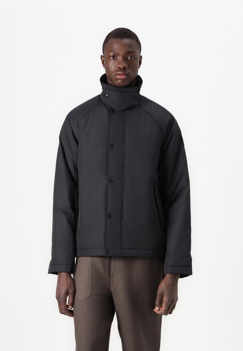 Black padded jacket with a high collar, snap-button closure, and side pockets. Features a smooth texture and minimalistic design.