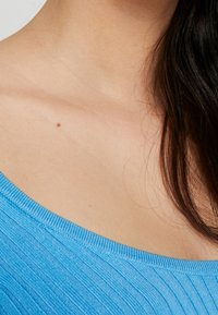 Niebieski prążkowany dekolt swetra z odsłoniętymi ramionami. Widoczna miękka tekstura, ukazująca jasny odcień skóry z małą pieprzykiem.