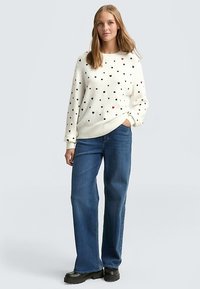 Pull en tricot crème avec des motifs de cœurs noirs et rouges, associé à un jean droit bleu et des bottes à plateforme noires. Encolure ronde simple.