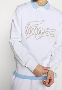 Sweat-shirt blanc avec un col bleu clair. Présente un logo détaillé de crocodile beige et un ourlet côtelé avec des accents bleus aux poignets.