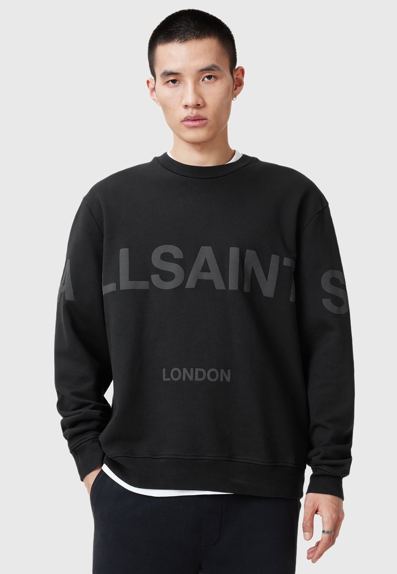 Jeune homme portant un sweat à capuche noir avec le texte "ALLSAINTS LONDON", se tenant devant un fond gris clair uni.