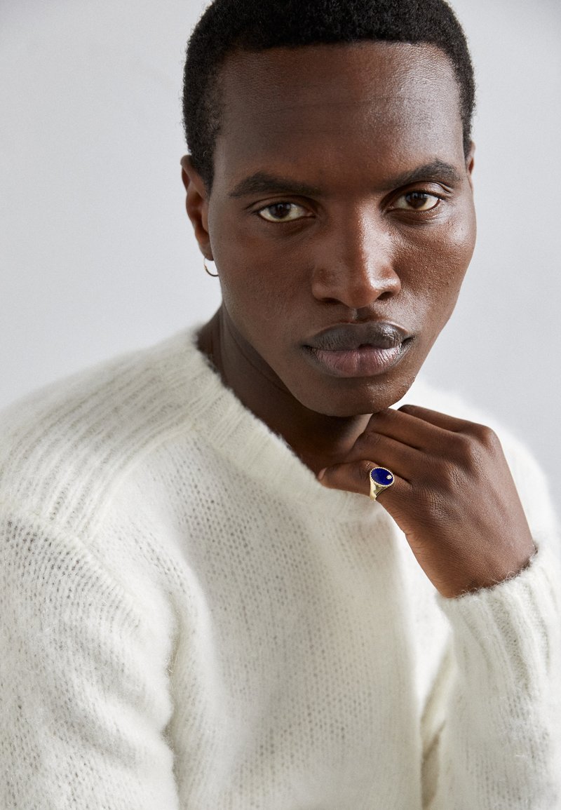 Bílý pletený svetr s volným střihem. Modelka má na pravé ruce zlatý prsten s velkým modrým kamenem a výrazné rysy obličeje.