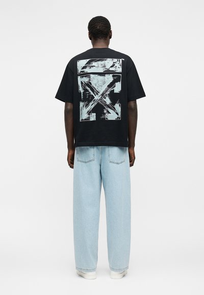 Άτομο που φοράει μαύρο μπλουζάκι με μεγάλο άσπρο αφηρημένο σχέδιο βέλους στην πλάτη, ανοιχτό μπλε τζιν παντελόνι με φαρδιά πόδια και λευκά αθλητικά παπούτσια.