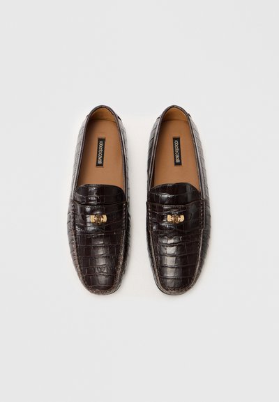 Ett par mörkbruna loafers i läder med krokodilsmönster, guld-tonade detaljer och beige sula på en vit bakgrund.