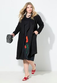 Schwarzer Wildleder-Mantel mit Fronttaschen, über einem schwarzen Kleid mit roten Blumenakzenten geschichtet. Model trägt rote offene High Heels.