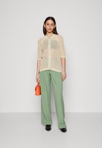 Beige mesh knappeskjorte med korte ermer, kombinert med løse grønne bukser og en oransje håndveske, står på en grå bakgrunn.