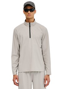Leichter, langärmeliger Pullover aus hellgrauem Stoff mit einem Viertel-Reißverschluss und einer strukturierten Oberfläche. Getragen mit passenden Hosen und einer Mütze.