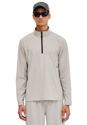 Leichter, langärmeliger Pullover aus hellgrauem Stoff mit einem Viertel-Reißverschluss und einer strukturierten Oberfläche. Getragen mit passenden Hosen und einer Mütze.