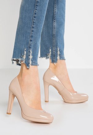 Beige laklæder højhælede sko med rundet tådesign og en 10 cm stiletthæl, kombineret med let slidte blå jeans.