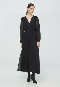 Abito lungo in chiffon nero con maniche lunghe trasparenti, scollo a V e vita marcata. Abbinato a stivali neri con tacco fino al ginocchio.