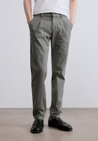 Επίπεδη, ανοιχτόγριζη chino παντελόνια με πλαϊνές τσέπες. Το ύφασμα φαίνεται λείο με ελαφριά υφή. Φορεμένα με μαύρα δερμάτινα παπούτσια.