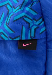 Μπλε σακίδιο πλάτης με γεωμετρικά σχέδια σε μπλε και τιρκουάζ, μαύρες μεταλλικές λεπτομέρειες και ροζ ετικέτα με το λογότυπο της Nike. Ανθεκτικό ύφασμα με υφή τελειώματος.