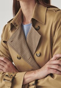 Béžový trench coat s límcem v károvaném vzoru a velkými knoflíky. Disponuje dvouřadým zapínáním a manžetami na přezku.