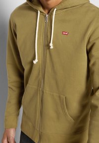 Felpa verde oliva con zip realizzata in tessuto morbido, dotata di una tasca frontale, cordini bianchi e un piccolo logo rosso Levi's sul petto.