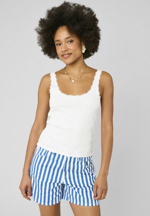 Femme aux cheveux bouclés portant un débardeur blanc avec bord festonné et un short à rayures verticales bleues et blanches, debout sur un fond uni.